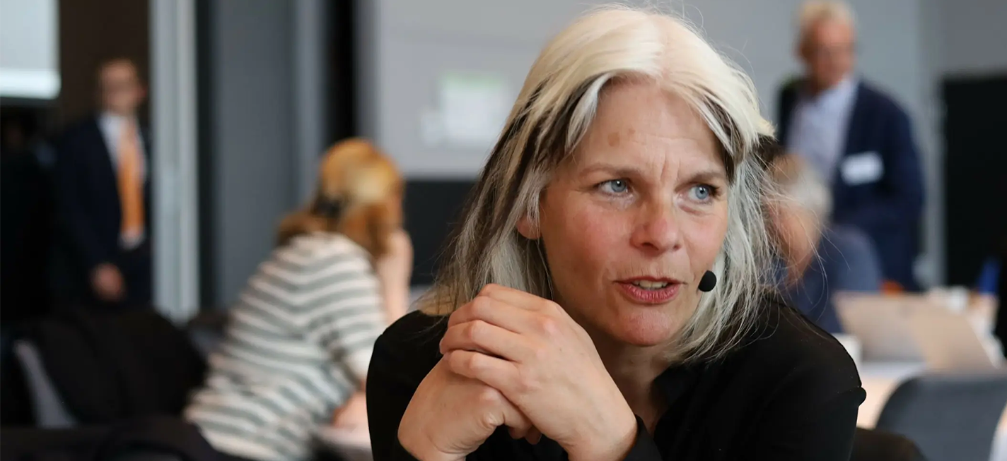 Professor Anne Kveim Lie talking in an academic setting with three figures in the background.
