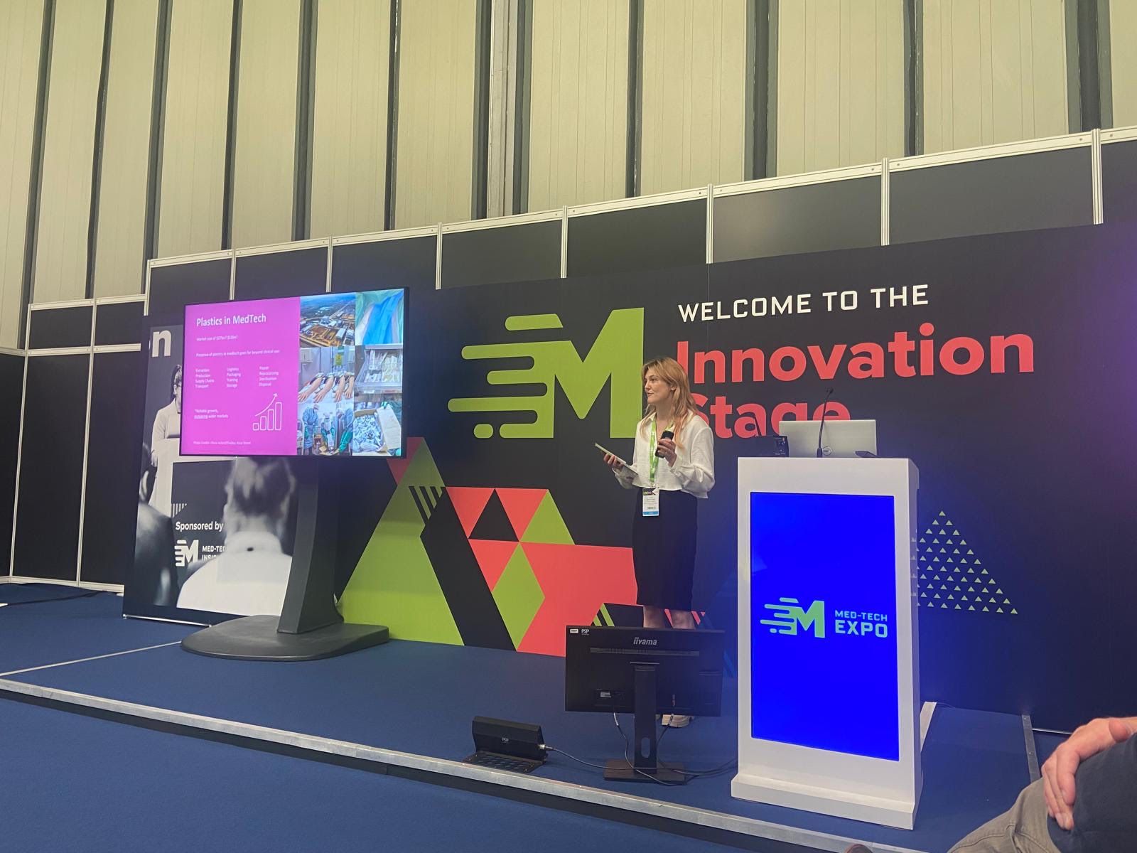 An exhibition presentation stage with screen and large dark coloured backdrop. In front is a researcher presenting with a microphone.