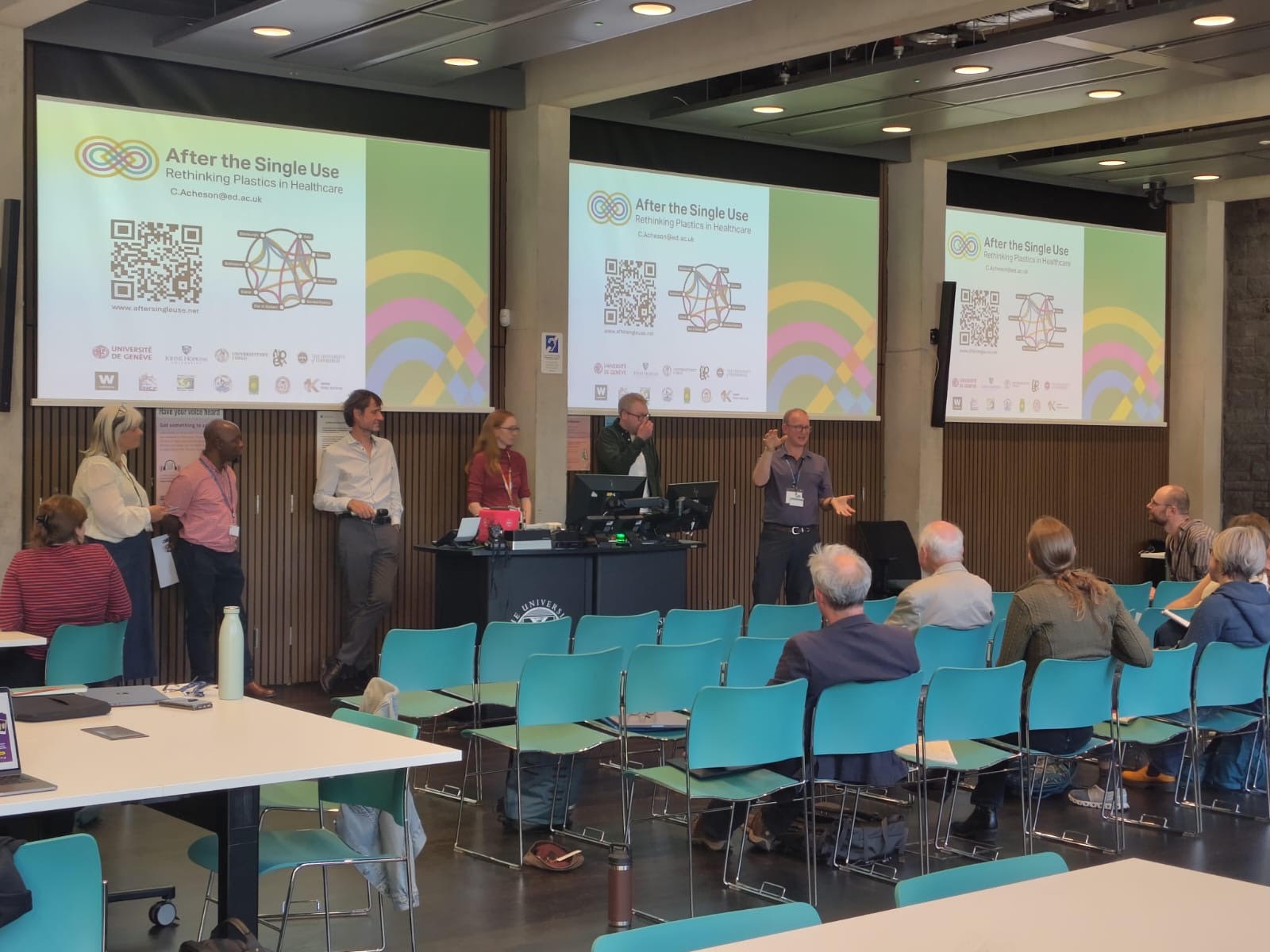 Researchers stand in front of 3 presentation screens in a room lined with tables and chairs.