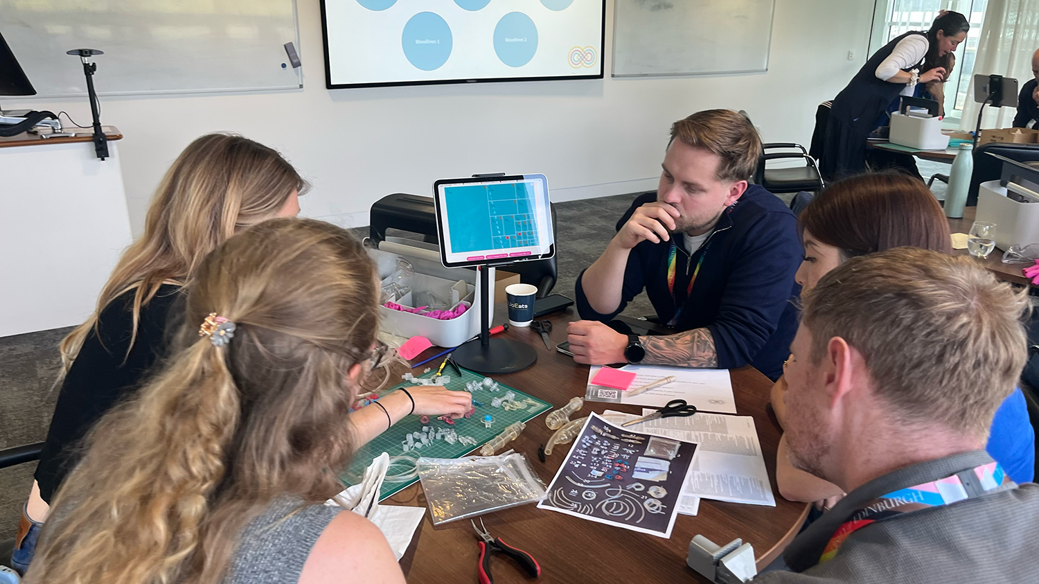 Workshop participants sit around a table with a tablet screen and a series of plastic components in front of them. 