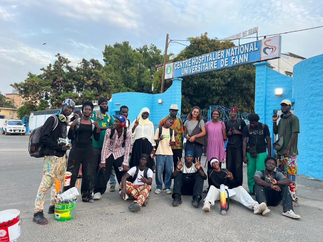 A group of people with paint speckled clothes stand in front of a blue graffiti wall.