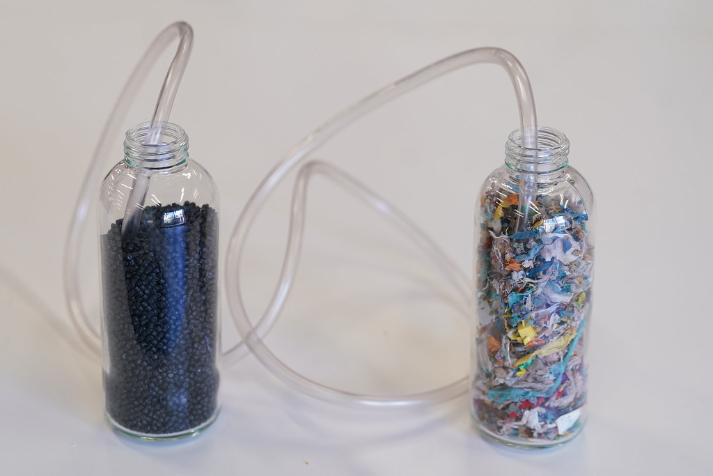 Two glass bottles, one containing small pastic pellets and another containing multicoloured shredded plastic, linked by a piece of medical tubing