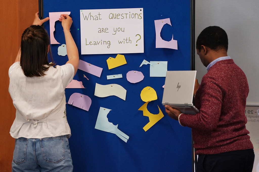 Anastasiia Martynenko and Amani Anaeli attaching coloured pieces of paper to a board with the text "What Questions are you Leaving With" written in the centre