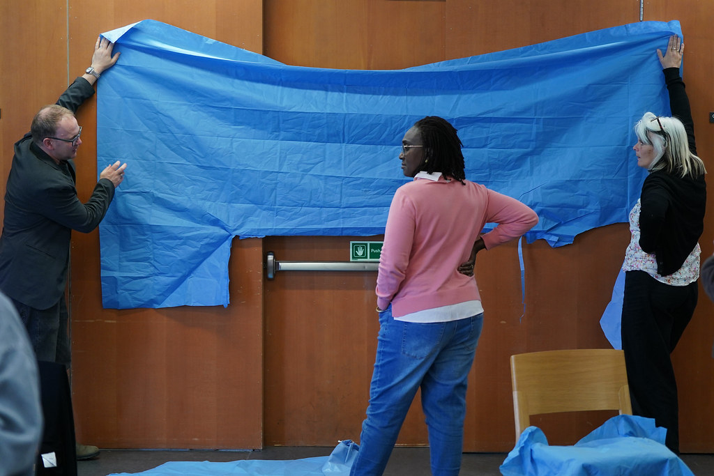 Two people holding each side of a large blue sheet of disposable surgical textile up against a wall with a third person in the middle