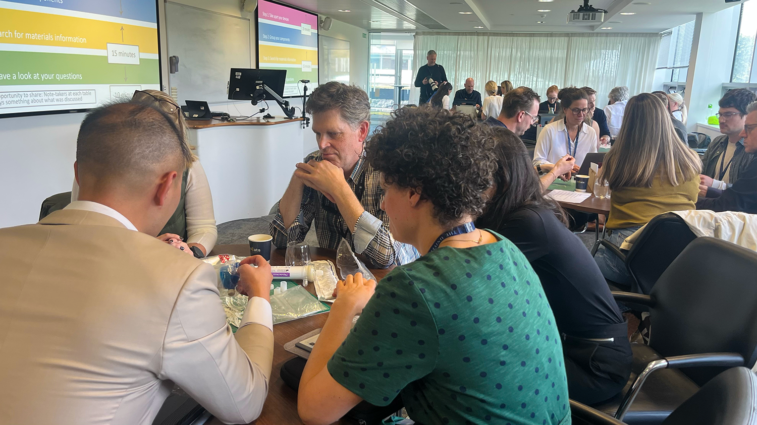 A meeting room filled with people around five tables looking thoughtfully at different plastic components and actively in discussion.
