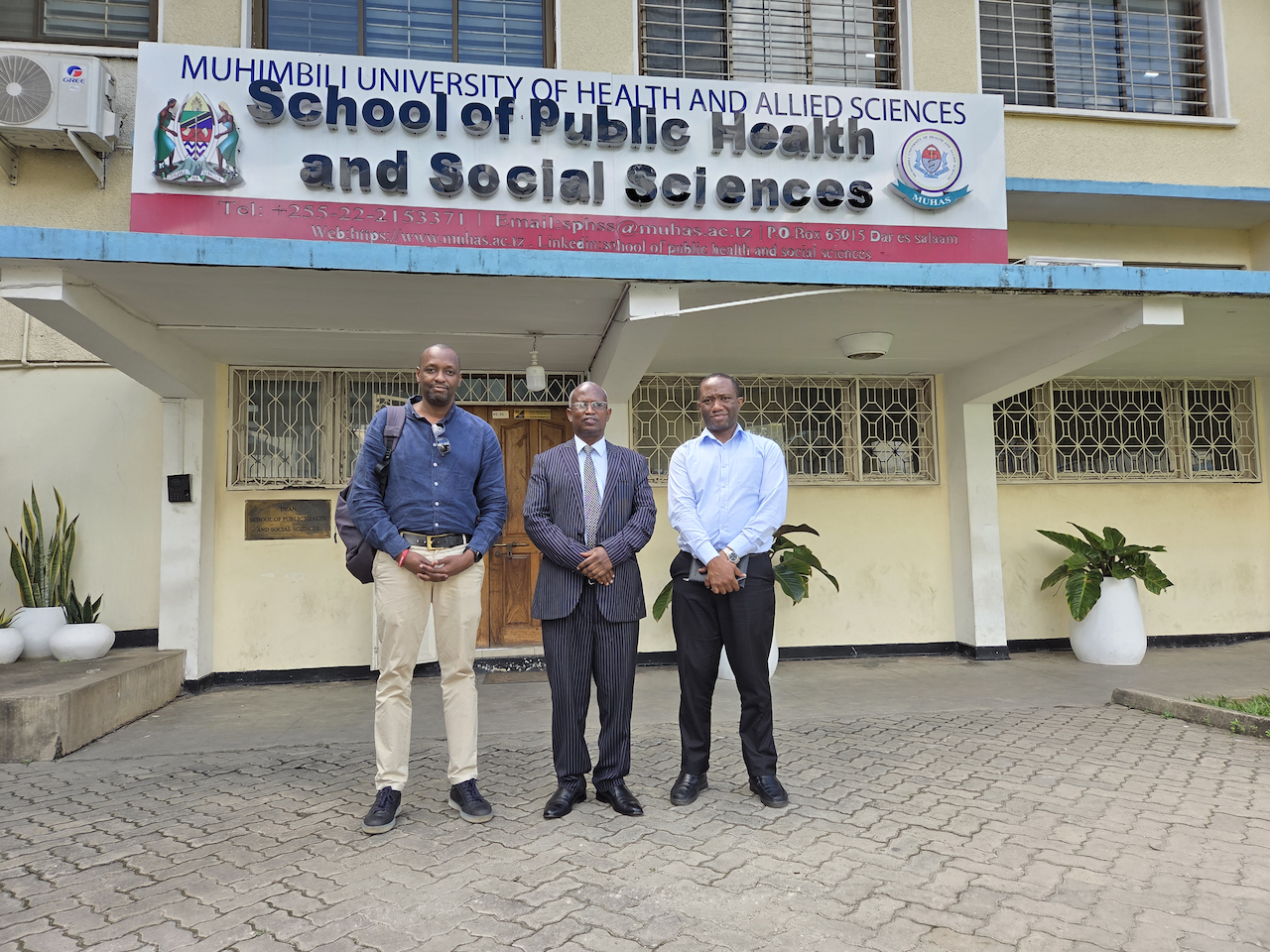 Researchers and representatives stand outside the School of Public Health and Social Sciences in Muhimbli. The building has a marigold plaster exterior with decorative metal work on the windows.