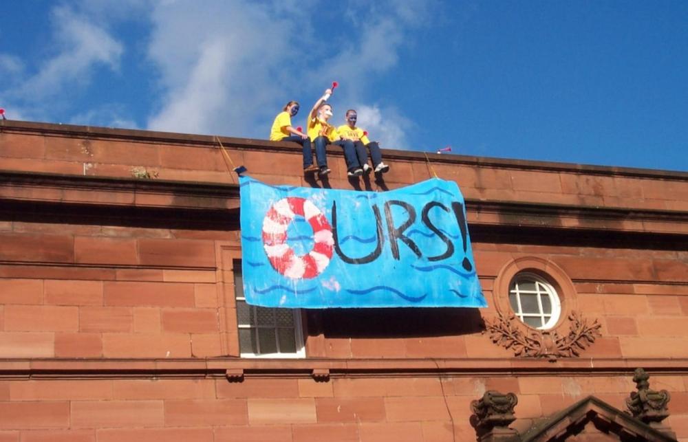 Three community activists occupy Govanhill baths, sitting on the roof of the baths with a banner that reads 'Ours' in 2001 when the pool was threatened with closure