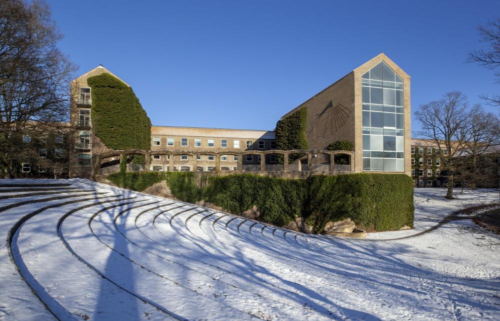 A large snow covered concourse with long steps foregrounds a modernist style university building with a glass fronted facade on one wing of the light brick building. 