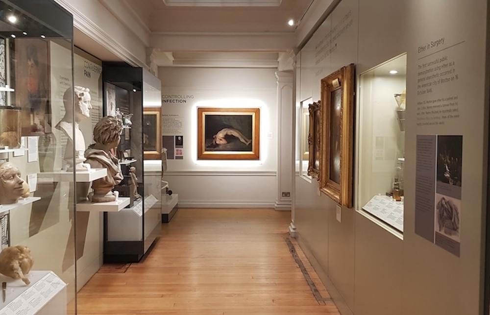 A series of displays line the muted white walls of the History of Surgery Museum. A oil painting with a thick frame is on the end wall. 