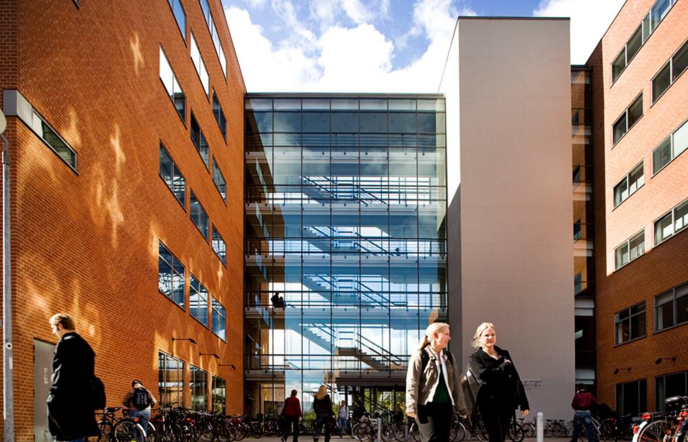 A brick and glass series of buildings at School of Culture and Society at Aarhus University are the backdrop for a bustling group of students and academics in the foreground. 