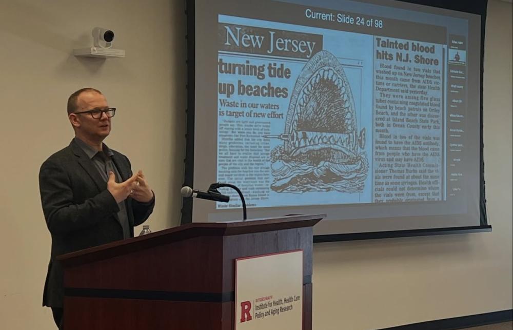 Jeremy Greene stands at a lecture in a suit with a slide on the projector showing a cartoon shark eating a syringe from a New Jersey press clipping. 