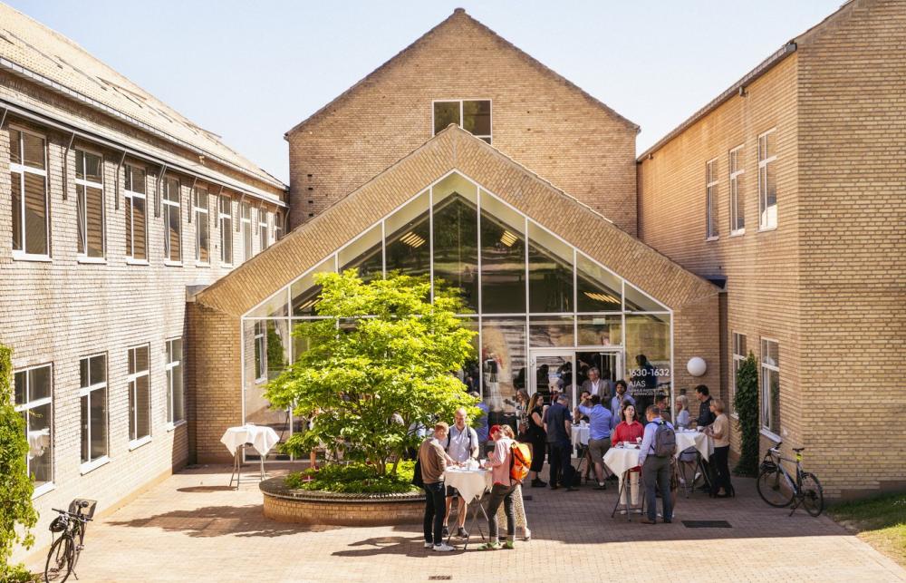 A group of people stand and chat outside of a peaked roof building with a glass front, there's a tree in the centre of the group in the courtyard. 