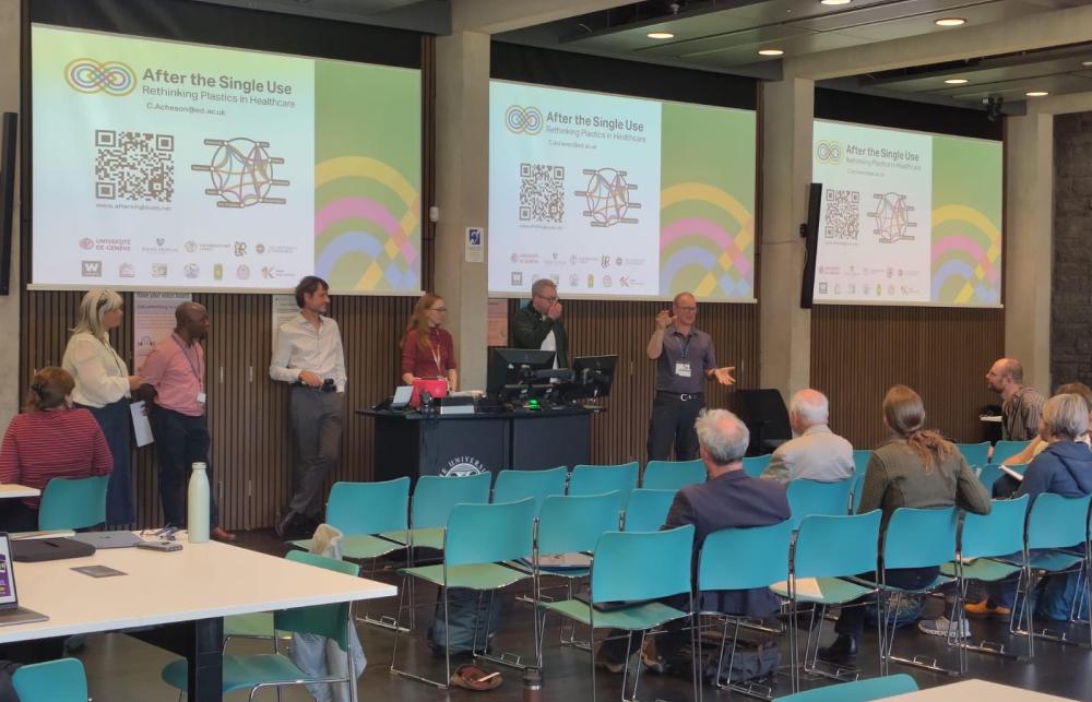 Researchers stand in front of 3 presentation screens in a room lined with tables and chairs. 